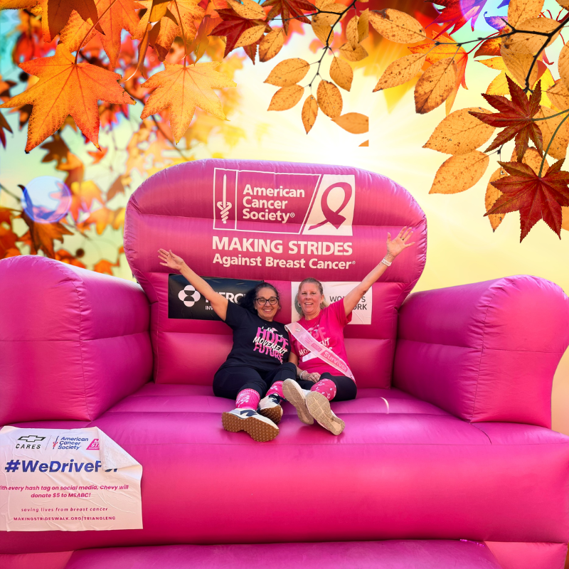 Two women sitting in the big pink inflatable chair with American Cancer Society branding against a colorful autumn leaf background for making strides against breast cancer