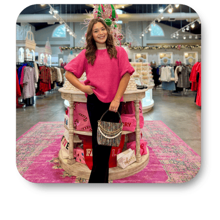 Woman in a pink sweater standing in the boutique with various clothing items at Dewayne's in Selma, NC minutes from Raleigh, Cary, Clayton, Garner, Smithfield, Goldsboro