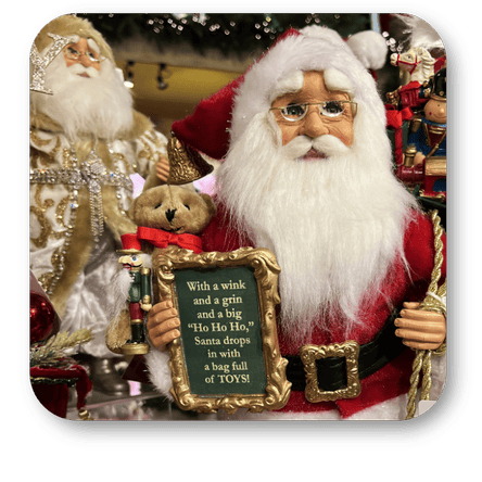 Decorative Karen Didion Santa Claus figure holding a sign with text, surrounded by Christmas-themed items in the timeless treasures room in christmas land at Dewayne's in Selma, NC minutes from Raleigh, Cary, Clayton, Garner, Smithfield, Goldsboro