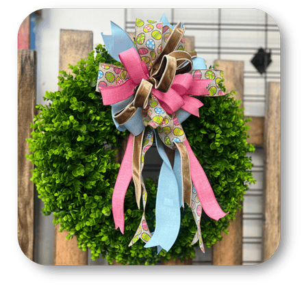 Green wreath with a colorful pastel happy bow on a wooden background Green wreath with a pink polka dot Happy bow to add the finishing touch in the greenhouse atrium at Dewayne's in Selma, NC minutes from Raleigh, Cary, Clayton, Garner, Smithfield, Goldsboro