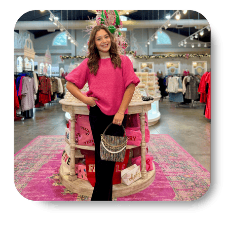 Woman in a pink sweater standing in the boutique with various clothing items at Dewayne's in Selma, NC minutes from Raleigh, Cary, Clayton, Garner, Smithfield, Goldsboro